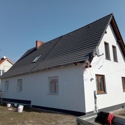 Świeżo otynkowana, biała elewacja domu jednorodzinnego z ciemną dachówką, widoczne zakryte okna i kilka wiader z farbą u podstawy ściany, sugerujące prace wykończeniowe.