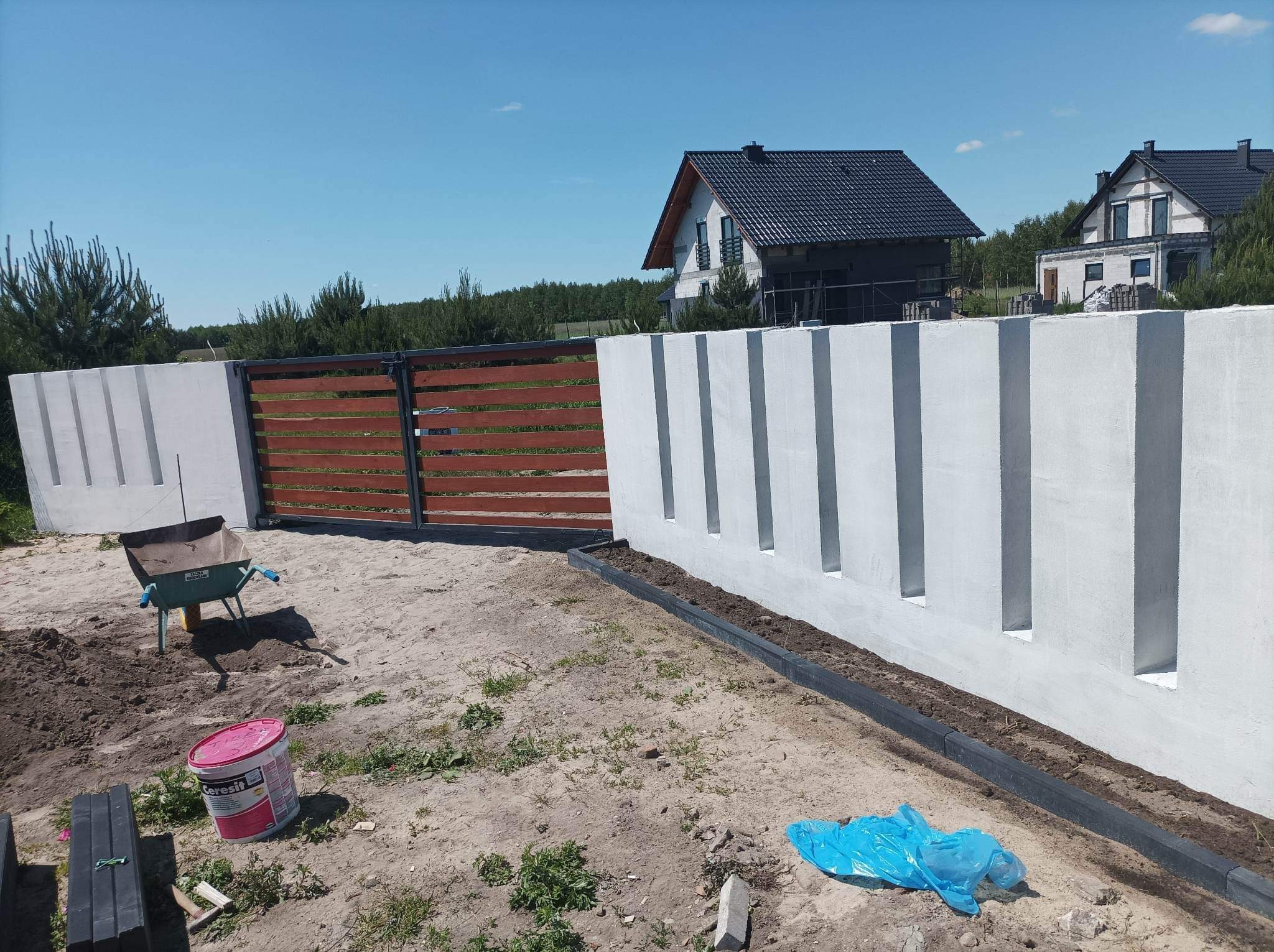 Nowoczesne ogrodzenie posesji z drewnianą bramą przesuwną i betonowym murem z dekoracyjnymi wnękami, widok na budynek w trakcie wykończenia, taczka i wiadro z zaprawą na pierwszym planie.