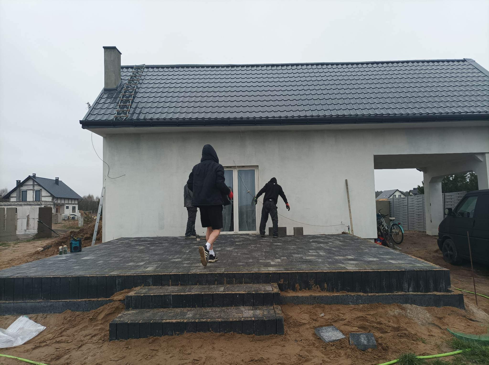 Czarny taras z kostki brukowej w trakcie budowy, z trzema schodami i pracownikami układającymi ostatnie elementy, widok na dom z szarą dachówką i samochód dostawczy.