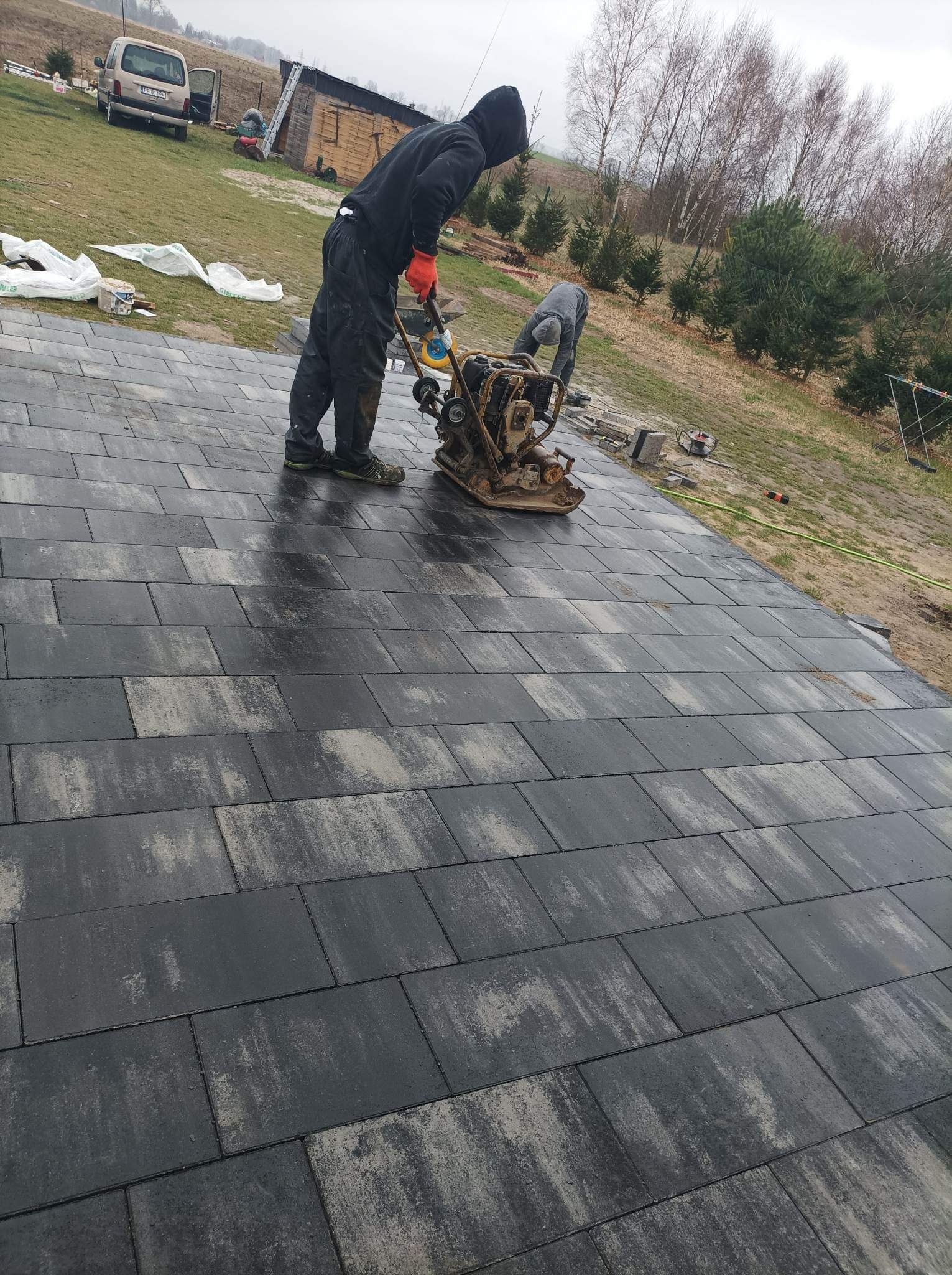 Pracownicy zagęszczają wibroprasą ułożoną, szaro-czarną kostkę brukową na posesji, widoczny samochód i budynek gospodarczy w tle.