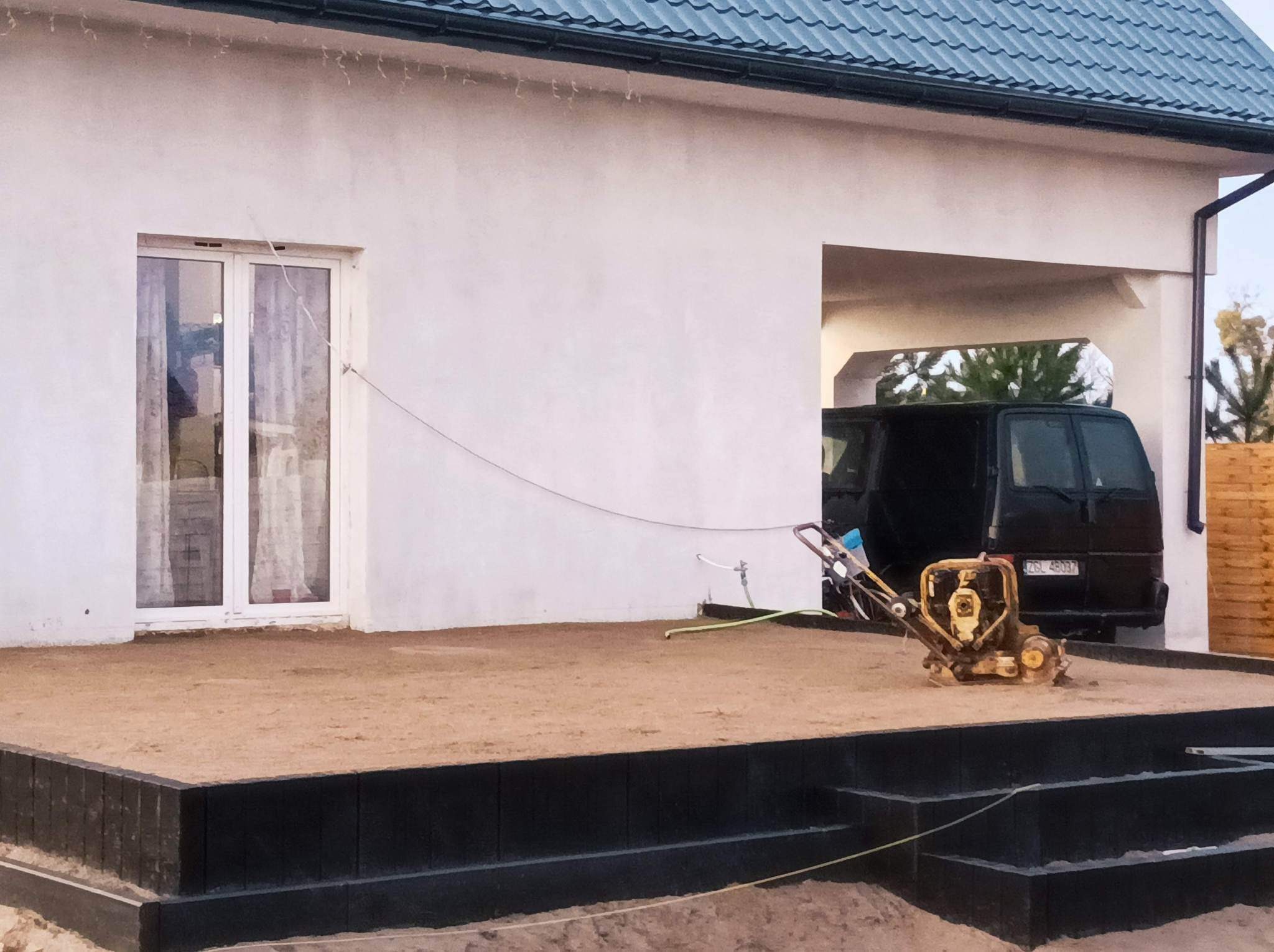 Przygotowanie podłoża pod taras: ubijarka wibracyjna na piasku, obrzeża z czarnych kostek, w tle biały dom z widocznym garażem i czarnym vanem.