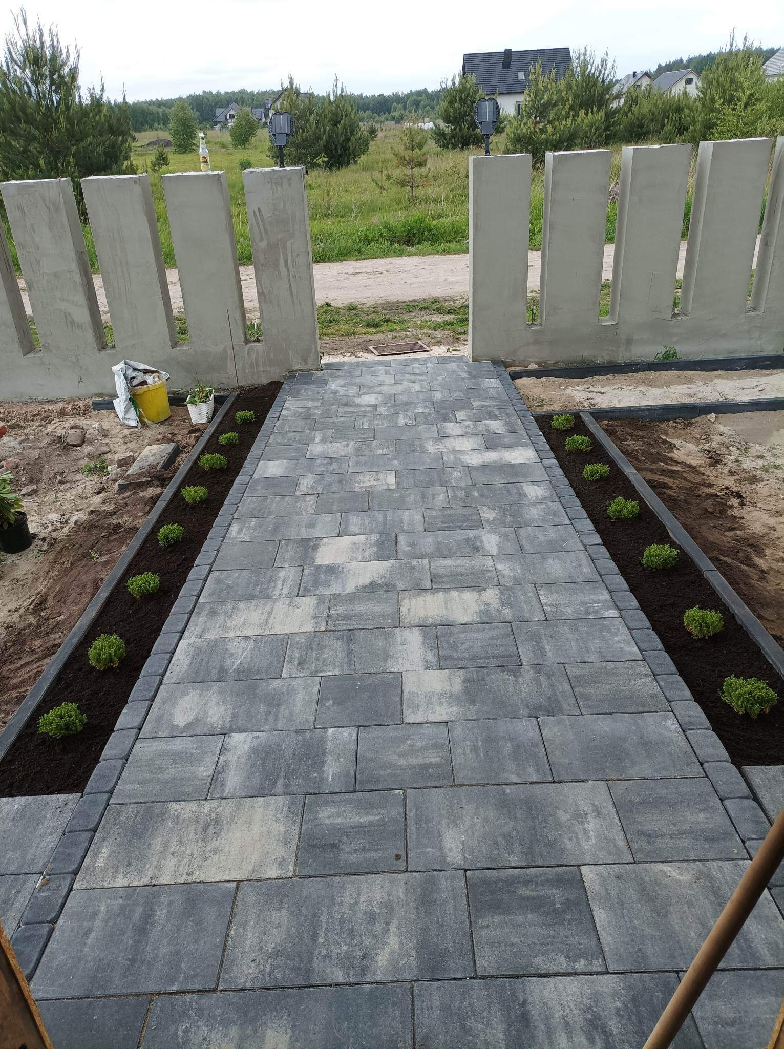 Ścieżka z szarej kostki brukowej prowadząca do betonowego ogrodzenia z charakterystycznymi prześwitami, obsadzona po bokach młodymi roślinami w świeżej ziemi.