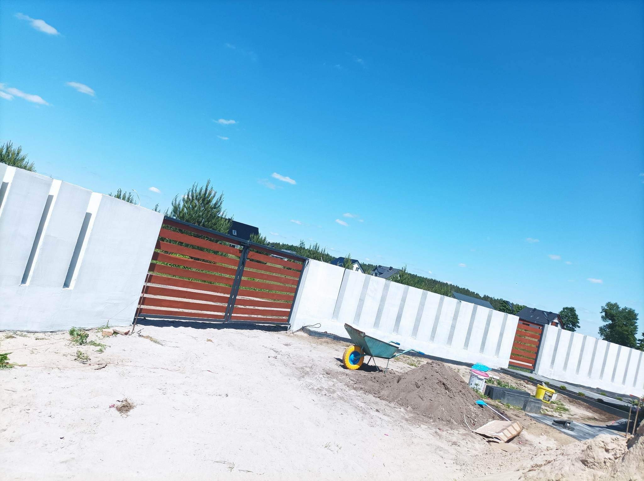 Nowoczesne ogrodzenie z białych betonowych przęseł i drewnianych paneli bramy przesuwnej, widok w trakcie prac ziemnych z taczką na pierwszym planie.
