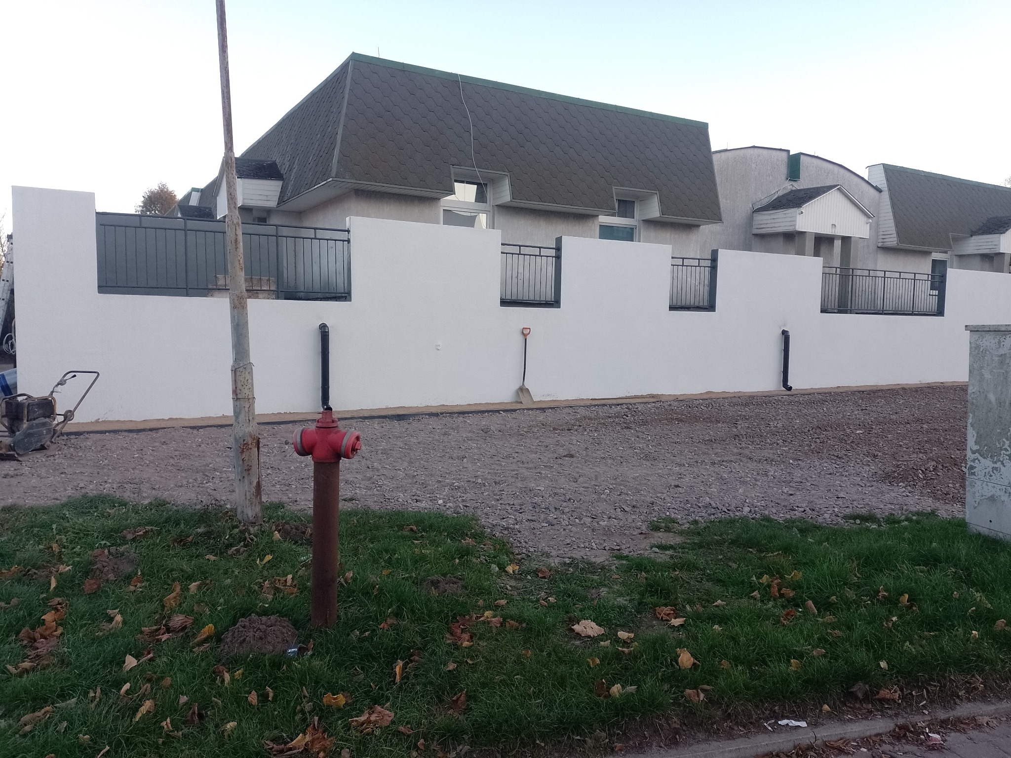 Świeżo otynkowany, biały mur z czarnymi, metalowymi przęsłami ogrodzeniowymi, widok na domy w tle, na pierwszym planie trawa, gruz i czerwony hydrant.