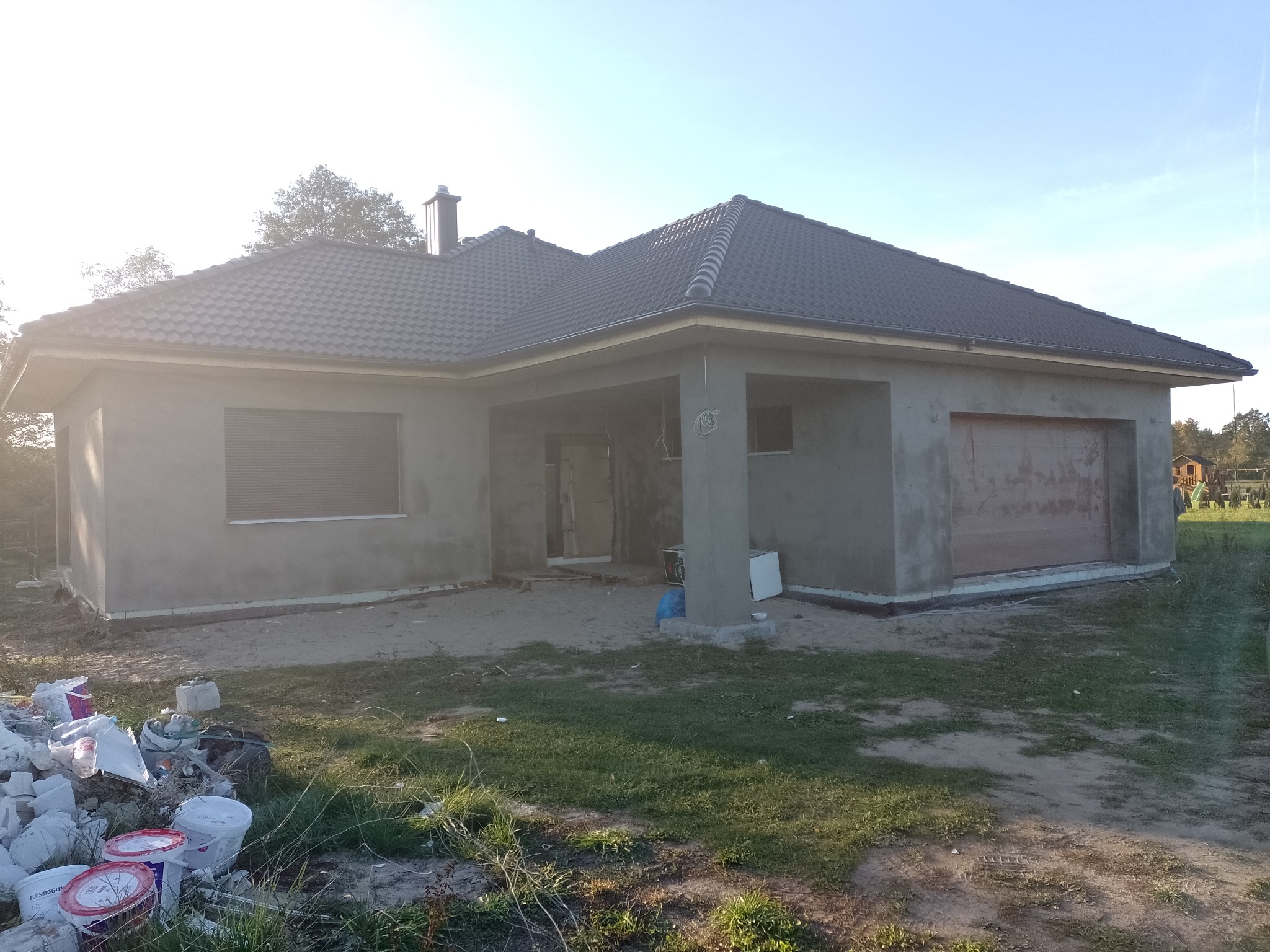 Surowa elewacja domu jednorodzinnego w trakcie prac wykończeniowych, widoczne puste wiadra po farbie i gruz, szare tynki i rolety w oknach, dach pokryty ciemną dachówką.