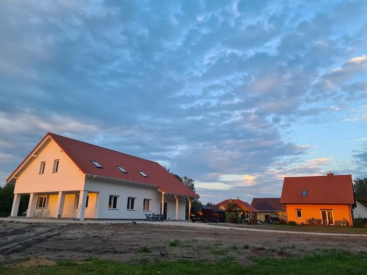 Nowo wybudowany, parterowy dom z białymi ścianami i czerwoną dachówką w otoczeniu innych budynków i terenu przygotowanego pod ogród, uchwycony w świetle zachodzącego słońca.