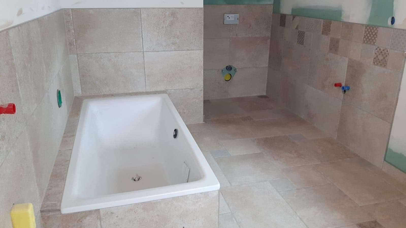 Wanna w łazience z beżowymi płytkami na ścianach i podłodze, częściowo wykończona, z widocznymi przyłączami wodnymi i fragmentem płyty gipsowo-kartonowej.