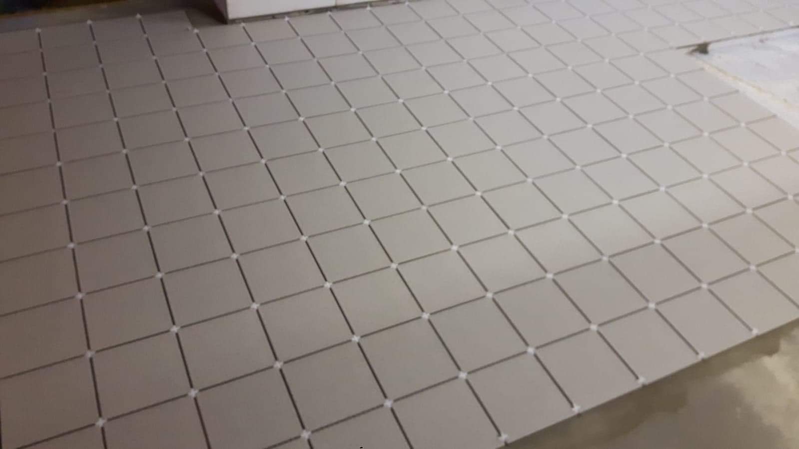 Ułożone, kwadratowe, matowe płytki podłogowe w odcieniach szarości z białymi krzyżykami dystansowymi, ułożone diagonalnie na surowym podłożu.