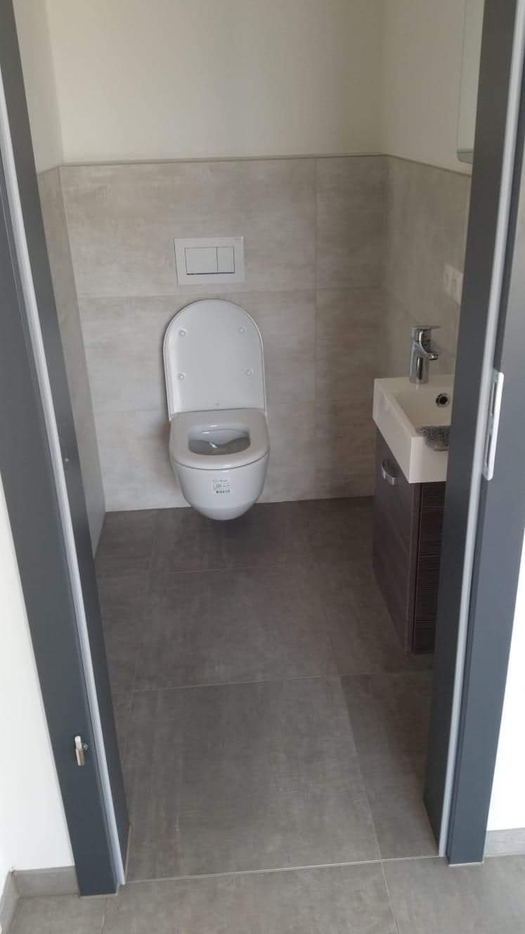 Wnętrze łazienki po remoncie: podwieszana toaleta z otwartą deską, umywalka z szafką w kolorze ciemnego drewna, szare płytki na ścianach i podłodze, widok zza szarej futryny drzwi.