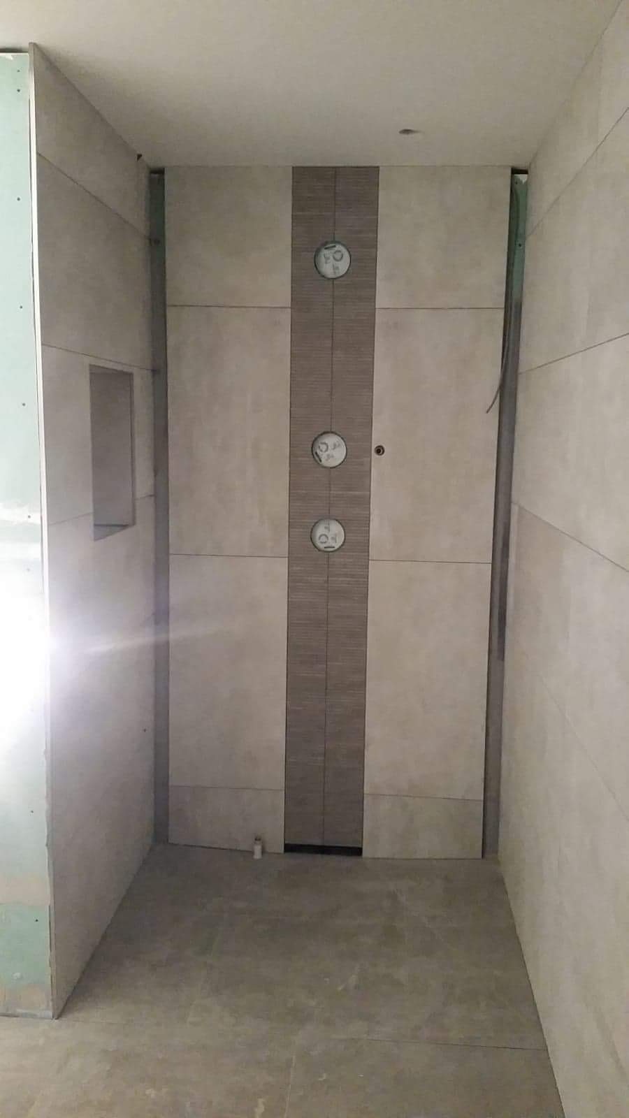 Wnętrze łazienki w trakcie wykańczania, widoczne płytki ścienne w odcieniach beżu z dekoracyjnym pasem z mozaiki, surowa ściana z płyt kartonowo-gipsowych oraz fragment podłogi z płytek.