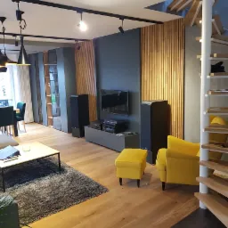 Nowoczesny salon z drewnianą podłogą, szarą ścianą z telewizorem, drewnianymi lamelami, spiralnymi schodami i żółtymi akcentami w postaci fotela i pufy.