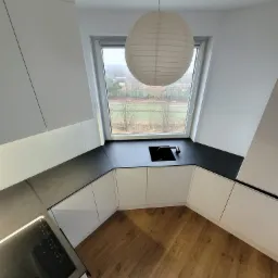 Nowoczesna kuchnia w kształcie litery U z białymi szafkami, czarnym blatem i drewnianą podłogą. Okno z widokiem na zewnątrz, nad blatem wisi okrągła lampa.