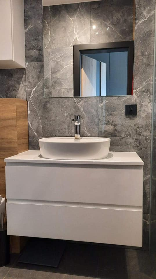 Nowoczesna łazienka z białą szafką pod umywalkę, okrągłą umywalką nablatową i baterią w kolorze chromu, na tle szarych płytek imitujących marmur oraz lustra w czarnej ramie.