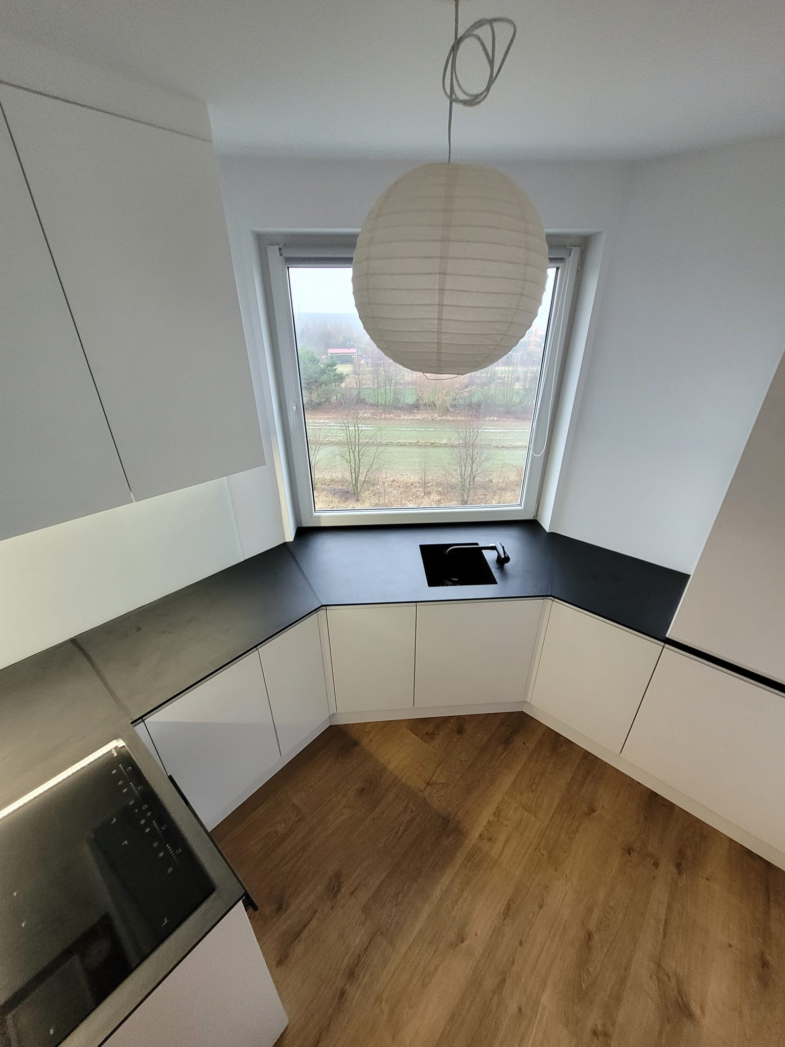 Nowoczesna kuchnia w kształcie litery U z białymi szafkami, czarnym blatem i drewnianą podłogą. Okno z widokiem na zewnątrz, nad blatem wisi okrągła lampa.