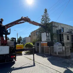 Praca dźwigiem Terex przy rozbudowie domu murowanego; robotnik w koszu dźwigu, blok z betonu komórkowego podnoszony w kierunku domu w trakcie rozbudowy, widoczne rusztowania.