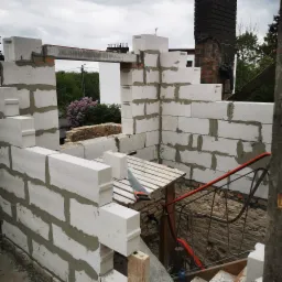 Wznoszenie ścian zewnętrznych domu z bloczków betonu komórkowego, widoczna piła ręczna na prowizorycznym podeście z desek, w tle mur z cegły i kwitnący krzew.