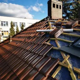 Montaż brązowej dachówki ceramicznej na drewnianej konstrukcji dachu, widoczny komin z cegły i budynek w tle pod błękitnym niebem.