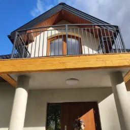 Dwukondygnacyjny dom z balkonem z drewnianą podłogą i metalową balustradą, wsparty na betonowych kolumnach, z drewnianymi drzwiami wejściowymi i oknem na balkonie.