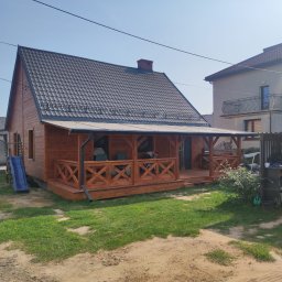 Usługi Stolarskie SOSENKA - Drewniany dom z ciemną dachówką i zadaszonym tarasem z balustradą w kształcie litery X. Obok domu zjeżdżalnia i huśtawka. W tle budynki mieszkalne i błękitne niebo.