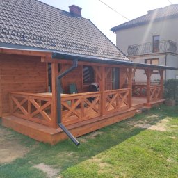 Usługi Stolarskie SOSENKA - Drewniany dom z zadaszonym tarasem z balustradą w kształcie krzyży. Widoczne rynny, dachówka i fragment ogrodu. Dom ocieplony, z widoczną strukturą drewna.