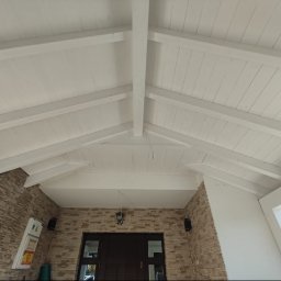 Usługi Stolarskie SOSENKA - Białe drewniane belki stropowe pod skośnym dachem, widok z dołu. Kamienna elewacja budynku z ciemnymi drzwiami wejściowymi. Oświetlenie zewnętrzne.