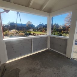 Usługi Stolarskie SOSENKA - Zadaszony taras z drewnianą, pomalowaną na szaro balustradą. Widok na ogród z domu jednorodzinnego w słoneczny dzień. Elementy konstrukcyjne w kolorze białym.