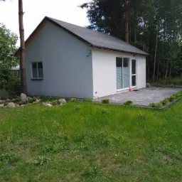 Niewielki, parterowy budynek z białymi ścianami i ciemnym dachem, otoczony zielonym trawnikiem i drzewami, z wybrukowanym tarasem przylegającym do ściany z przesuwnymi drzwiami.