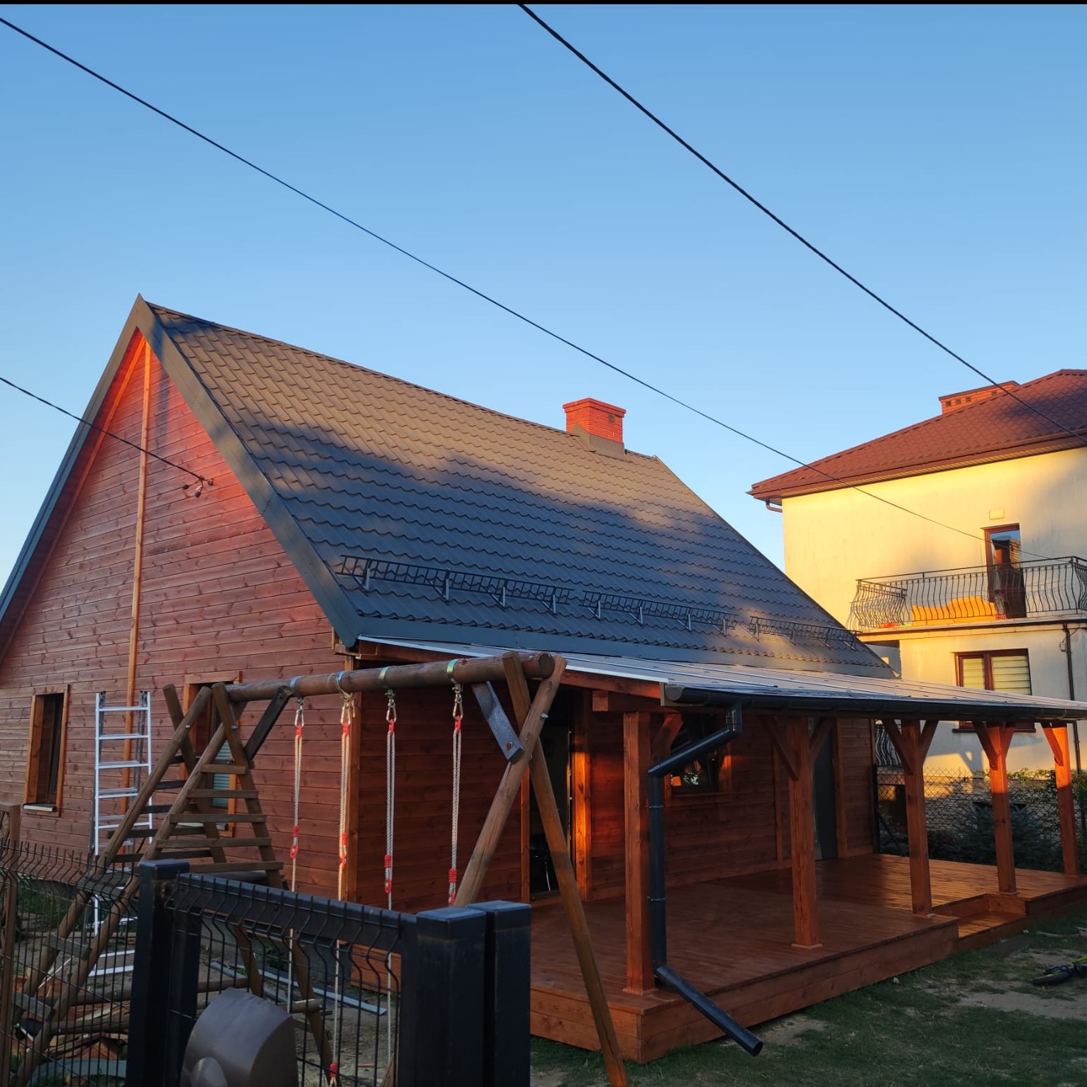 Drewniany dom z ciemnym dachem i zadaszonym tarasem. Obok huśtawka i drabina. W tle budynek mieszkalny. Ujęcie w słoneczny dzień.