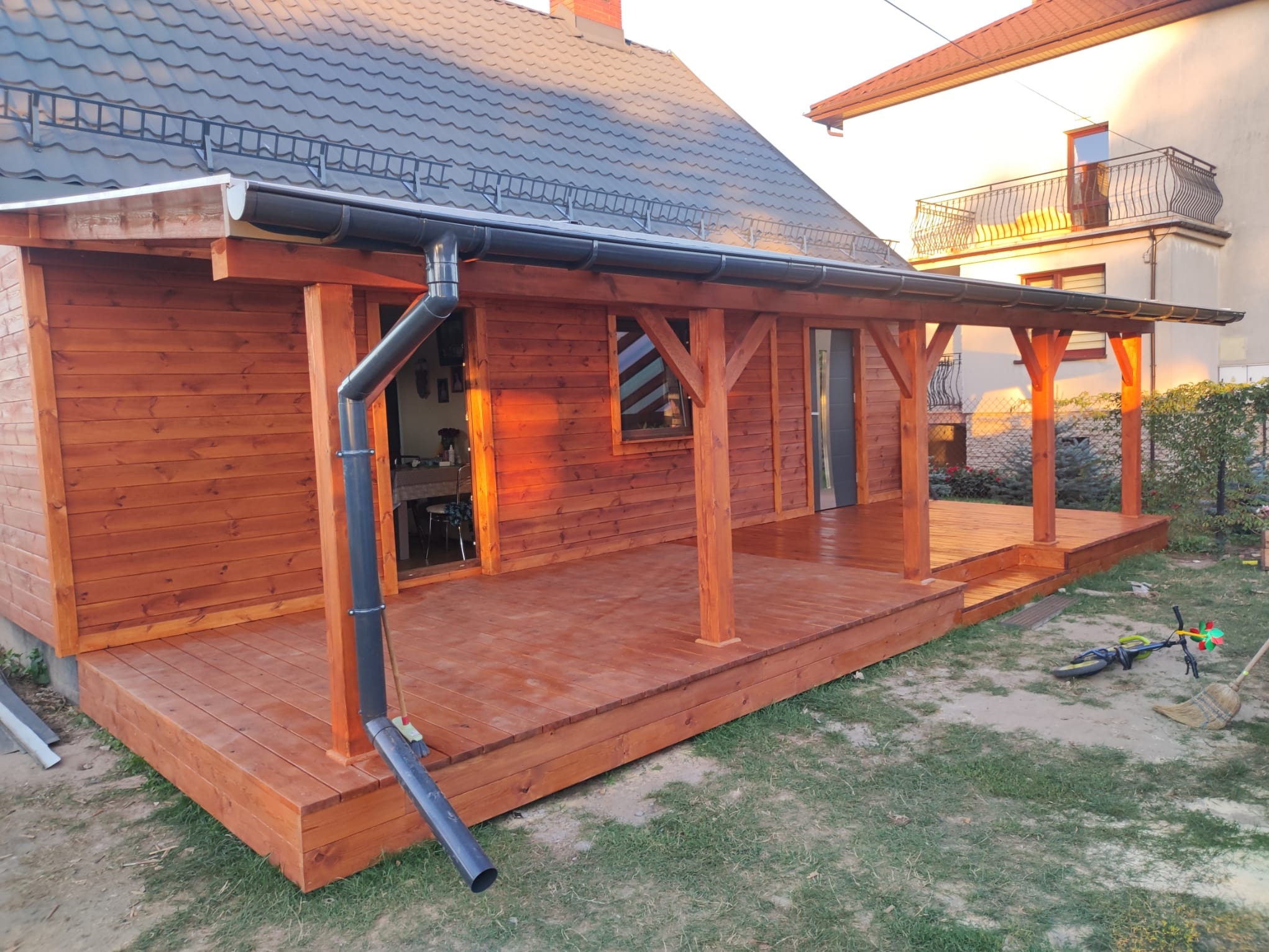 Drewniana konstrukcja z zadaszeniem i tarasem, świeżo po impregnacji, z widocznym systemem odprowadzania wody. W tle dom z szarą dachówką i balkonem.