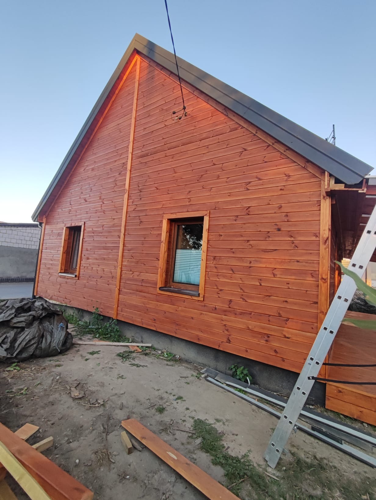 Zewnętrzny widok domu szkieletowego z drewnianą elewacją w ciepłym odcieniu brązu, z widocznymi dwoma oknami i fragmentem dachu. Na pierwszym planie drabina i teren budowy.