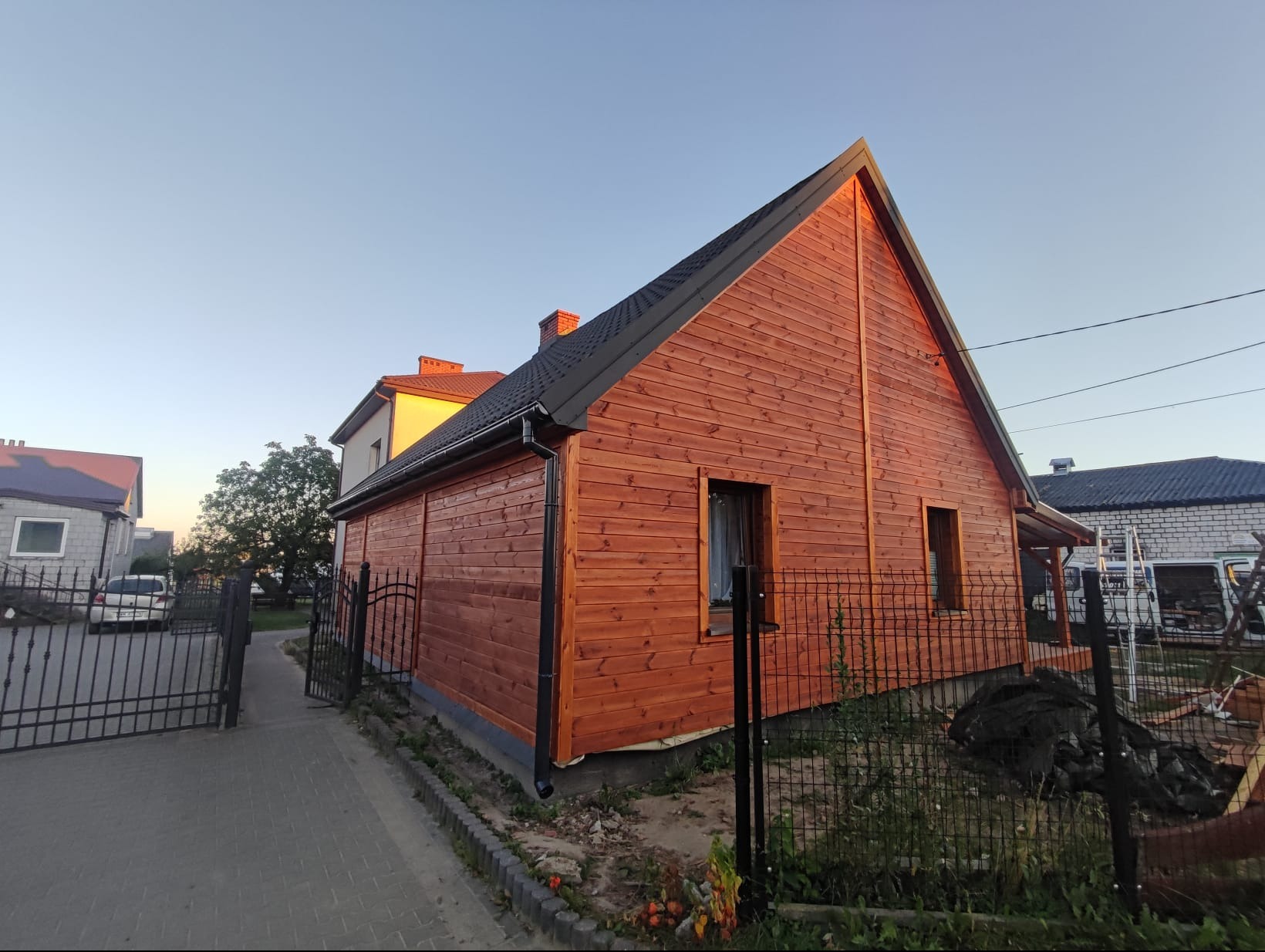 Ciepły odcień drewna na elewacji domu z dwuspadowym dachem, widok z boku, ogrodzenie z metalowych prętów, sąsiednie budynki w tle, słoneczny dzień.