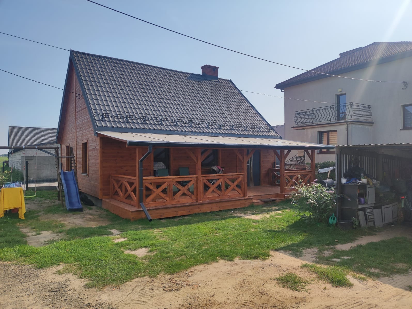 Drewniany dom z ciemną dachówką i zadaszonym tarasem z balustradą w kształcie litery X. Obok domu zjeżdżalnia i huśtawka. W tle budynki mieszkalne i błękitne niebo.