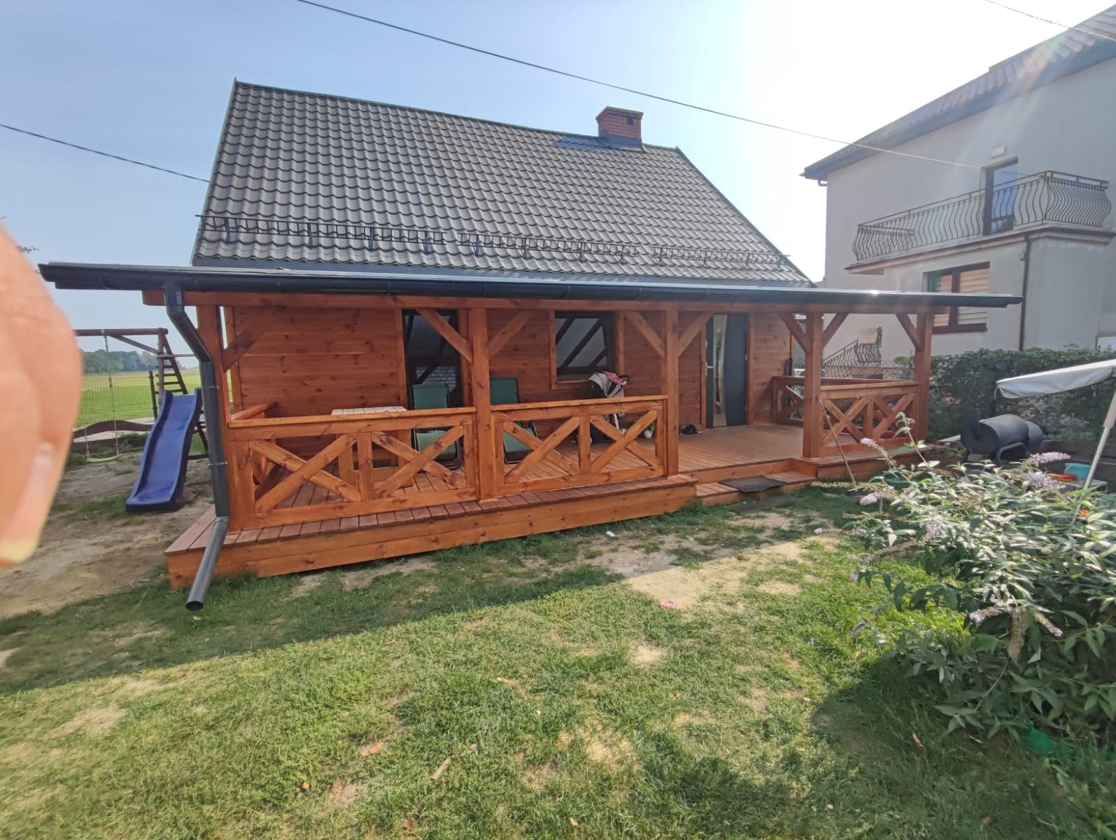 Drewniana konstrukcja z zadaszeniem i balustradą, prawdopodobnie domek letniskowy lub altana, z widocznym fragmentem zjeżdżalni i elementami otoczenia.