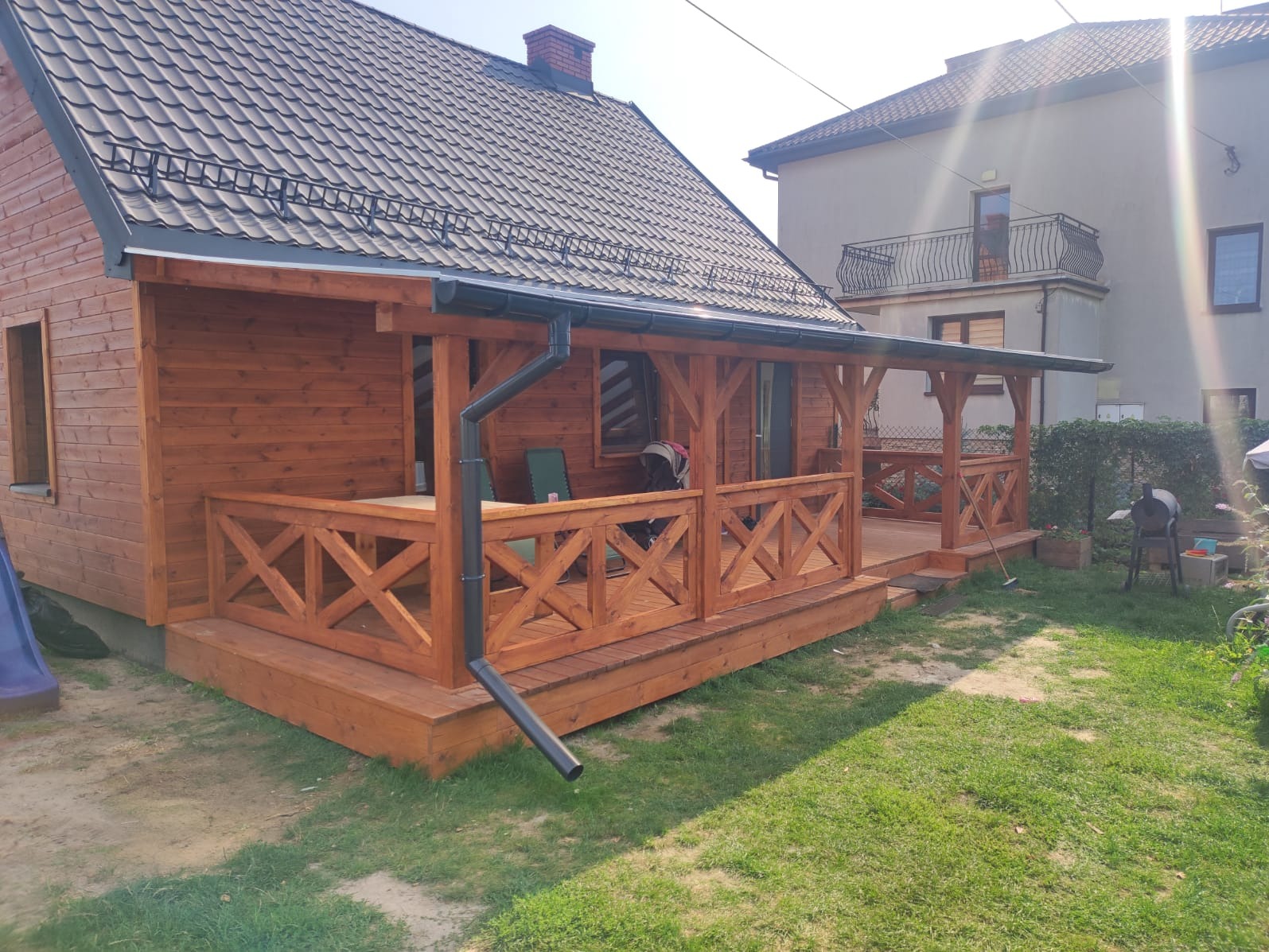 Drewniany dom z zadaszonym tarasem z balustradą w kształcie krzyży. Widoczne rynny, dachówka i fragment ogrodu. Dom ocieplony, z widoczną strukturą drewna.