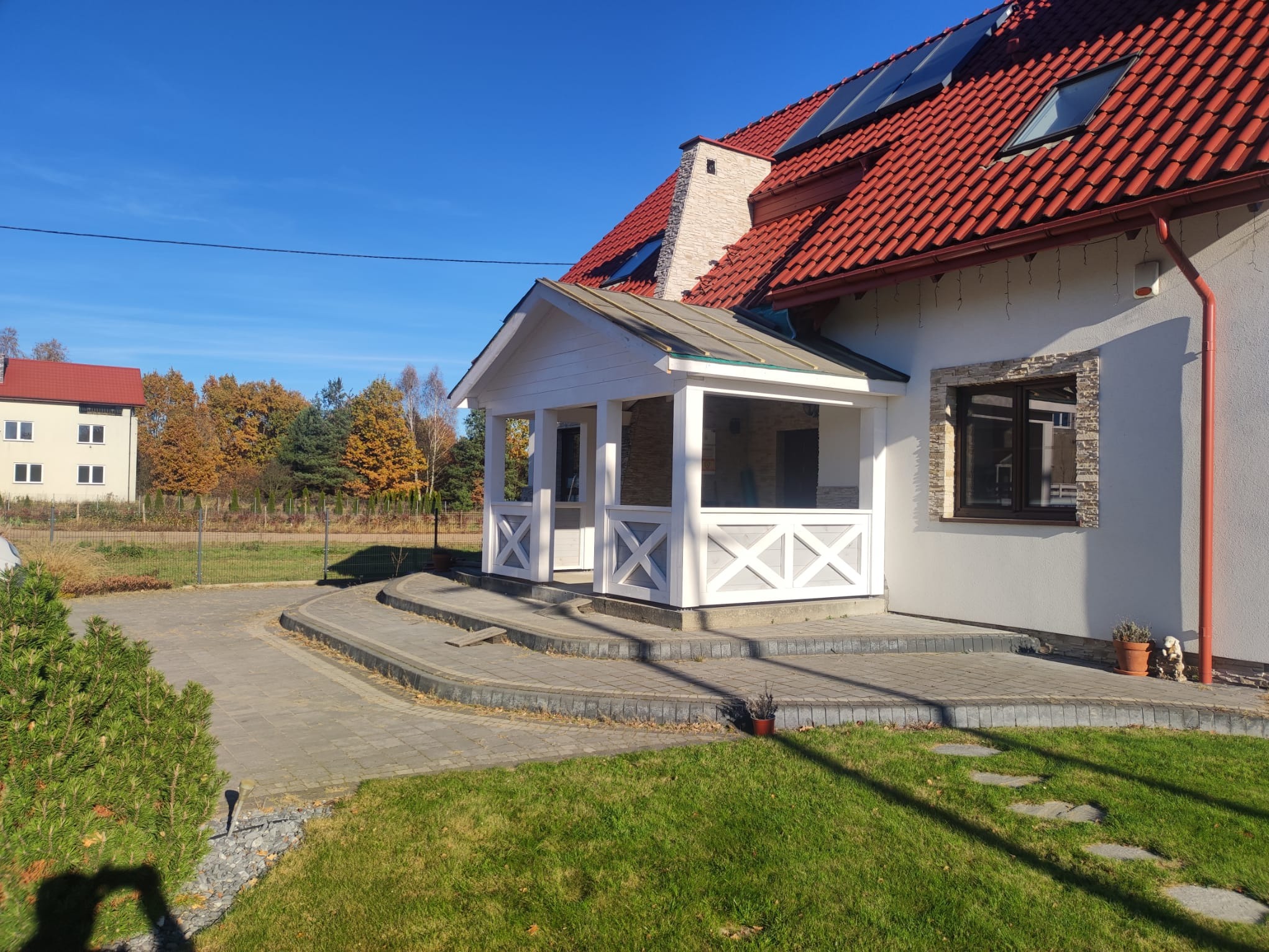 Biała, zadaszona weranda z dekoracyjnymi krzyżami, przylegająca do jasnego domu z czerwoną dachówką. Widok na zadbany trawnik i fragment ścieżki z kostki.