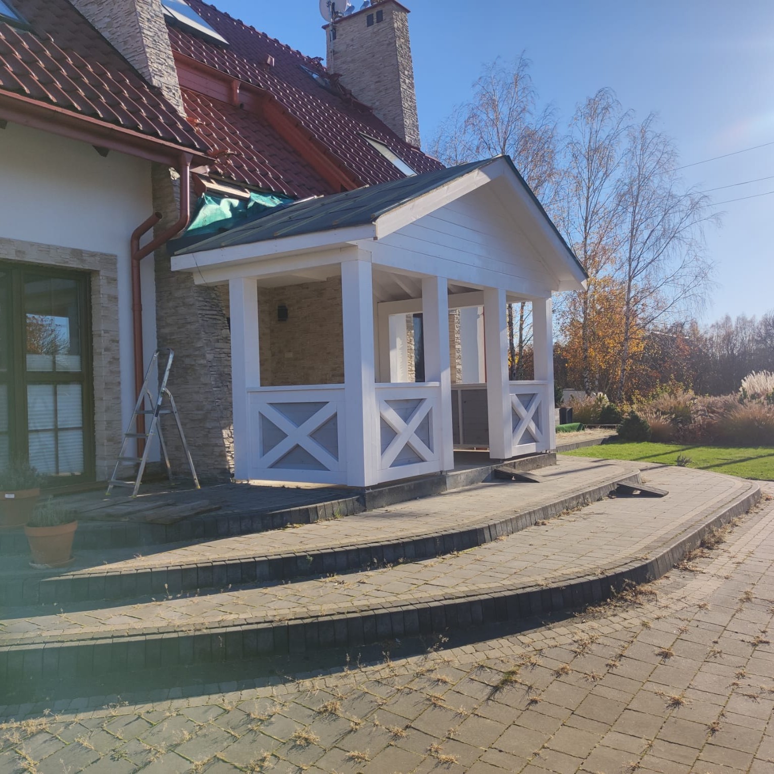 Biała, drewniana altana z dekoracyjnymi krzyżami, częściowo zadaszona, na kamiennych schodach przy domu z ceglanym dachem. W tle drzewa i trawa.