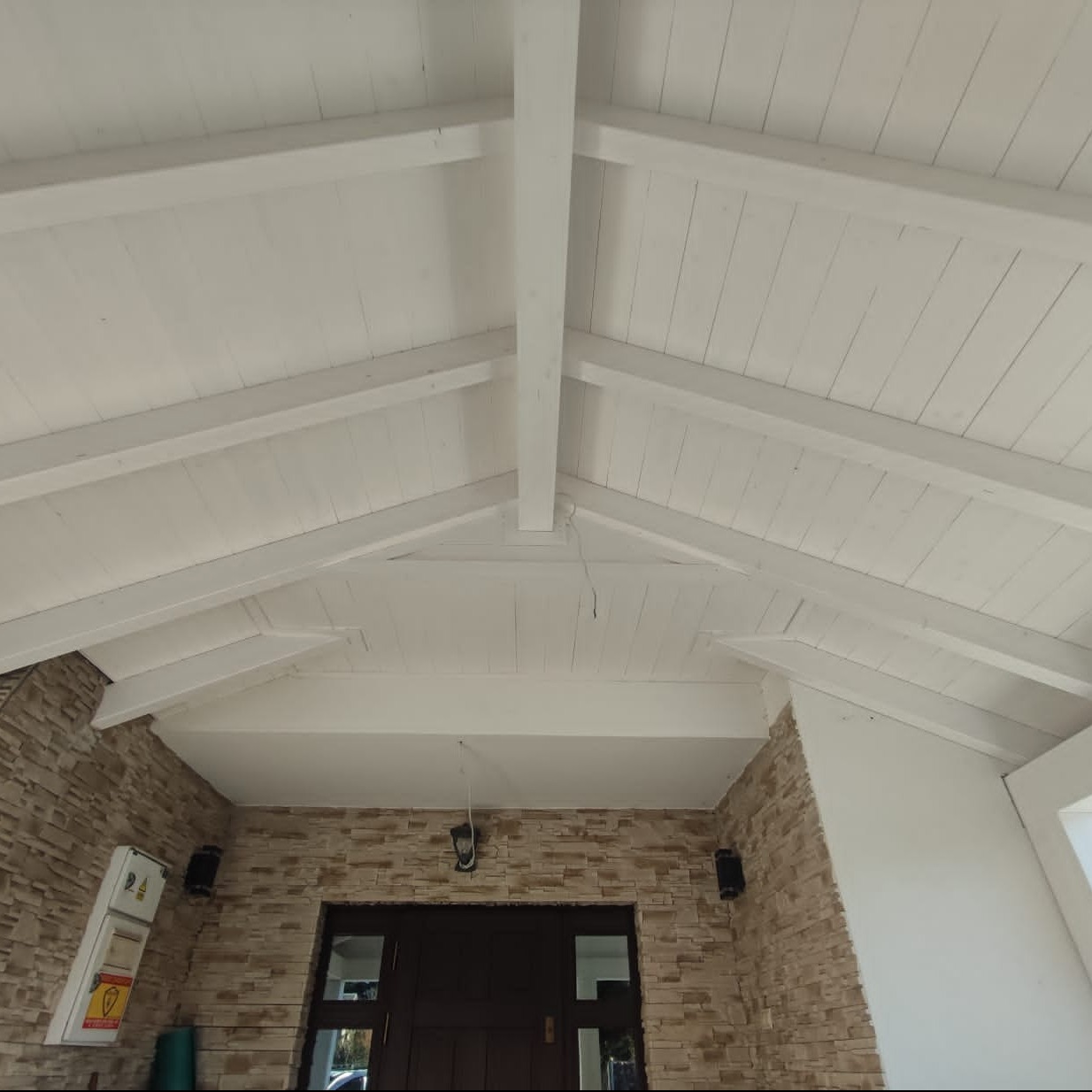 Białe drewniane belki stropowe pod skośnym dachem, widok z dołu. Kamienna elewacja budynku z ciemnymi drzwiami wejściowymi. Oświetlenie zewnętrzne.
