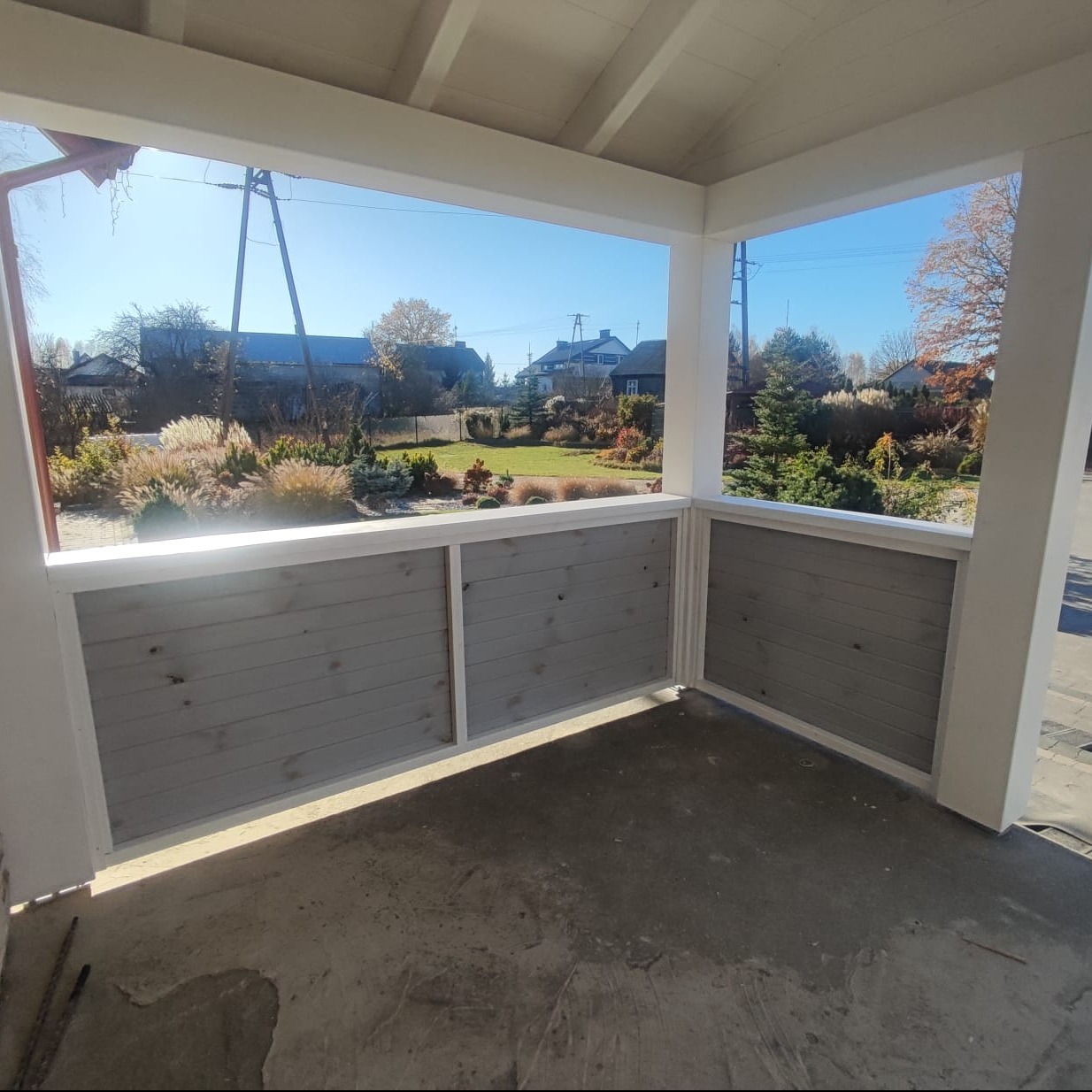 Zadaszony taras z drewnianą, pomalowaną na szaro balustradą. Widok na ogród z domu jednorodzinnego w słoneczny dzień. Elementy konstrukcyjne w kolorze białym.