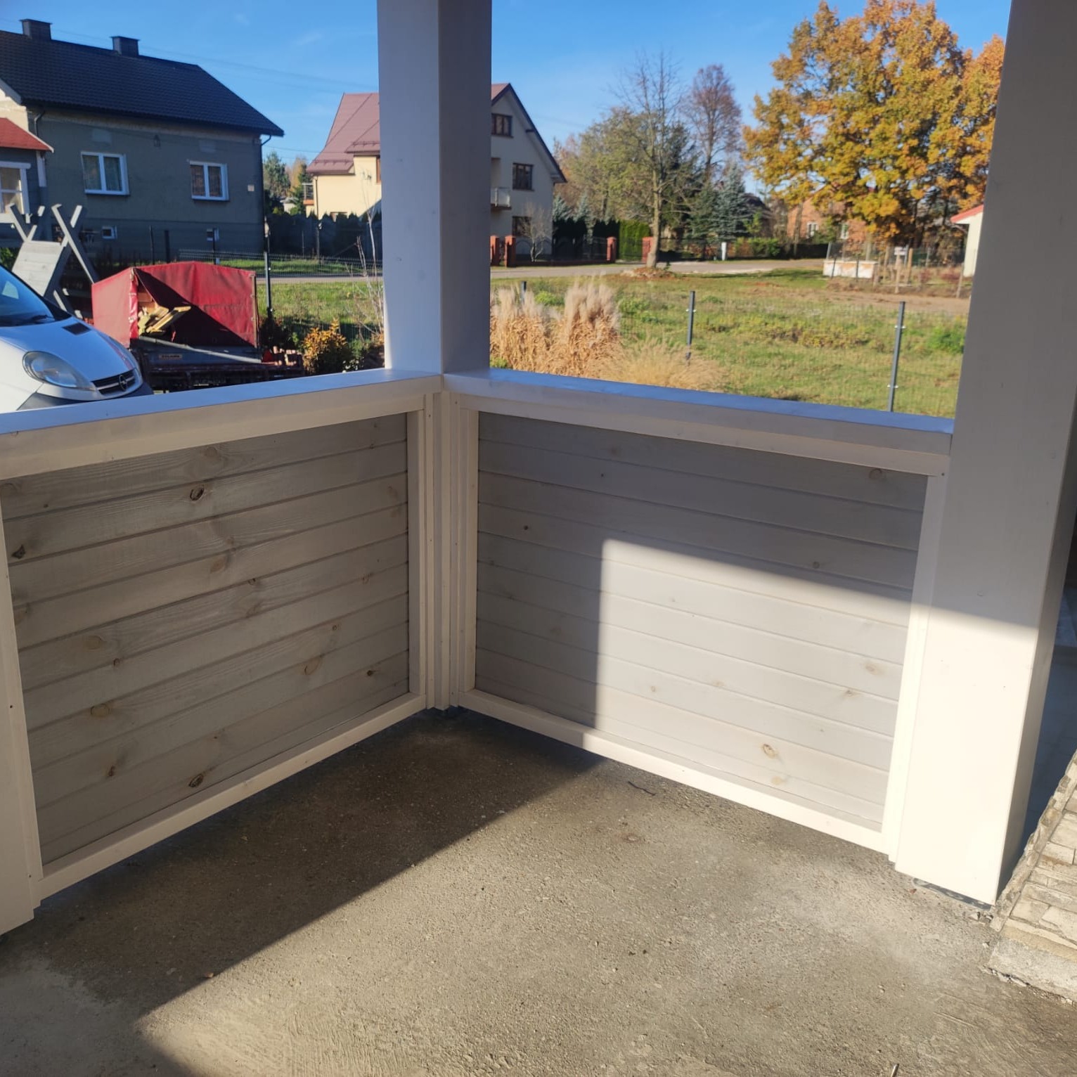 Wykończony balkon z drewnianą, pomalowaną na szaro boazerią i białymi słupami. Widok na ogród i zabudowę jednorodzinną w tle. Słoneczny dzień.