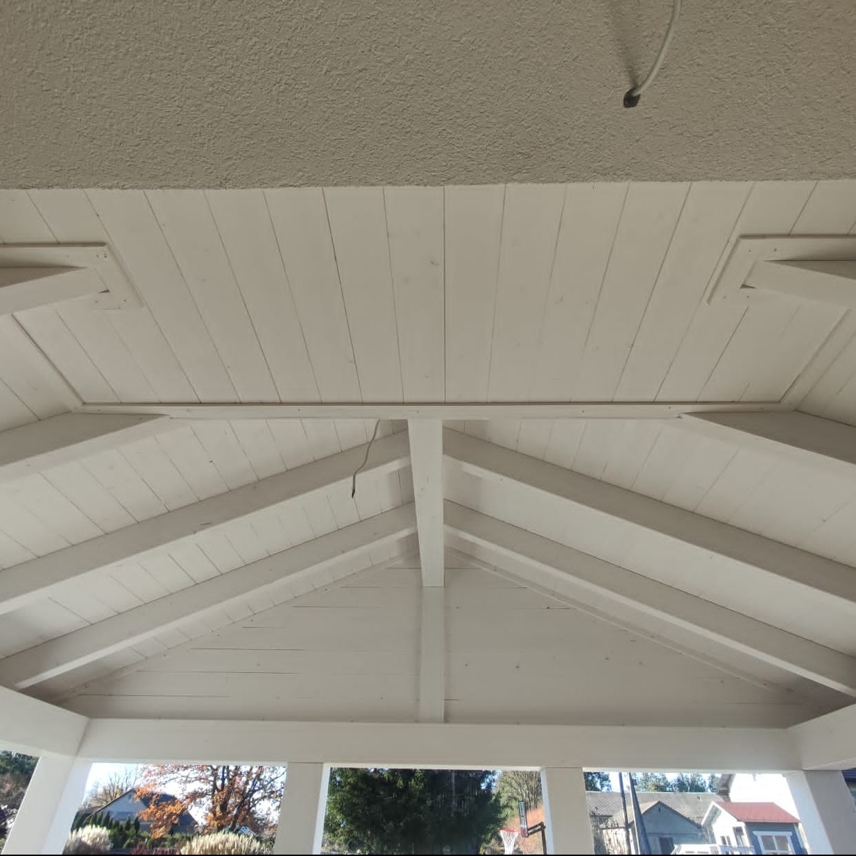 Biała, drewniana konstrukcja z widocznym dachem i belkami, widziana od wewnątrz. W tle zieleń drzew i zabudowa mieszkalna. Ujęcie z dołu.