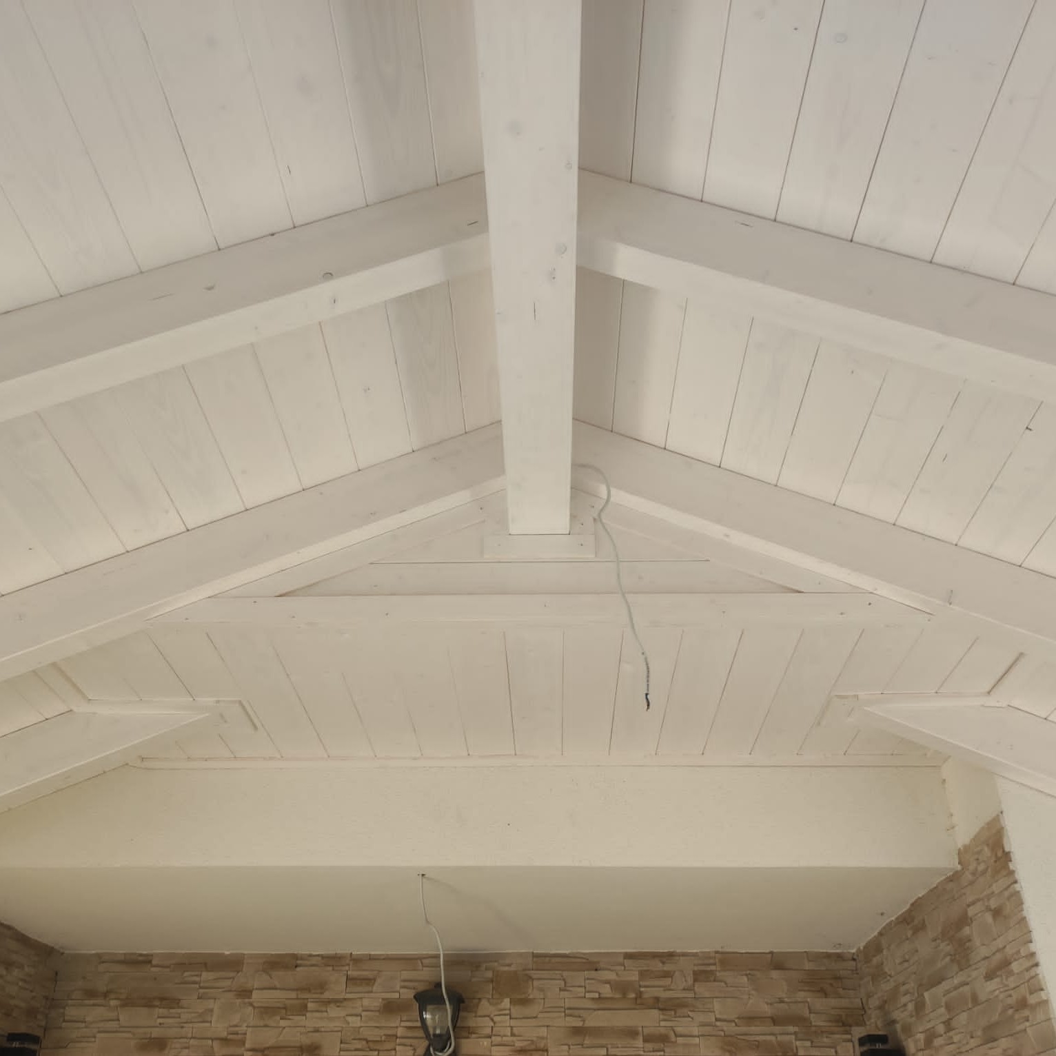 Białe drewniane belki stropowe tworzące trójkątny wzór, widok z dołu, z fragmentem ściany z dekoracyjnej cegły w dolnej części kadru.