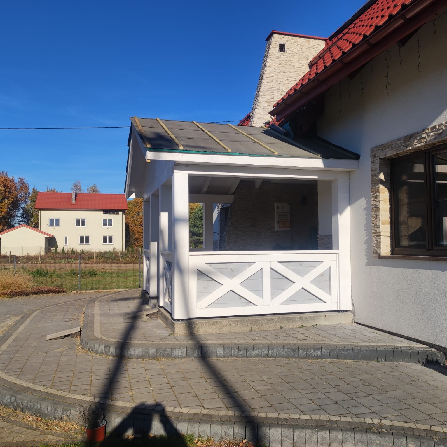 Biała, drewniana konstrukcja z dekoracyjnymi krzyżami, dobudowana do domu z czerwonym dachem. Widok z zewnątrz, na pierwszym planie kostka brukowa.