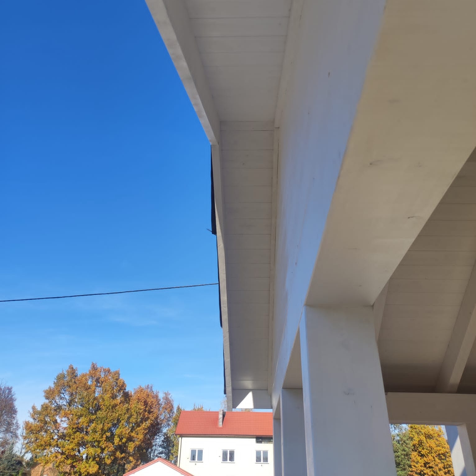 Błękitne niebo nad białą konstrukcją drewnianą z widocznymi belkami i fragmentem budynku z czerwoną dachówką w tle, jesienne drzewo po lewej stronie.