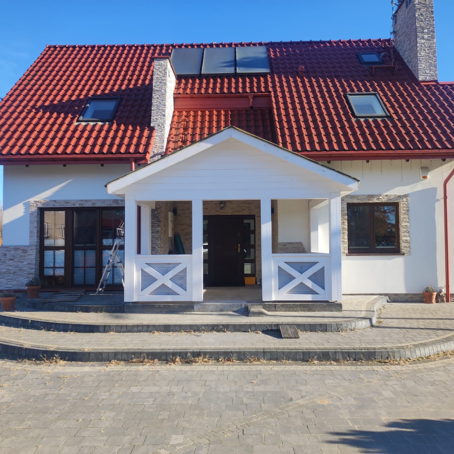 Biała, drewniana weranda z dekoracyjnymi krzyżami, dobudowana do domu z czerwoną dachówką i panelami słonecznymi. Schody z kostki brukowej prowadzą do wejścia.