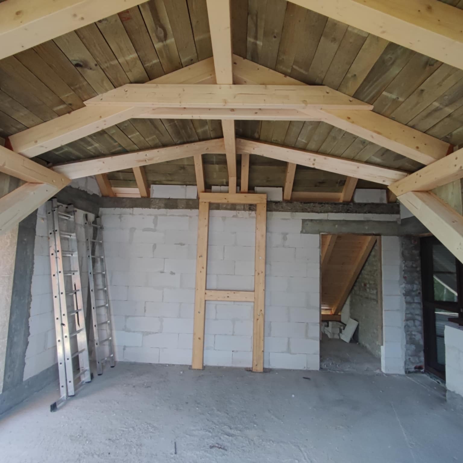 Wnętrze w trakcie budowy z drewnianą konstrukcją dachu i ścianami z bloczków. Widoczna drabina i drewniane ramy. Surowy stan wykończenia.