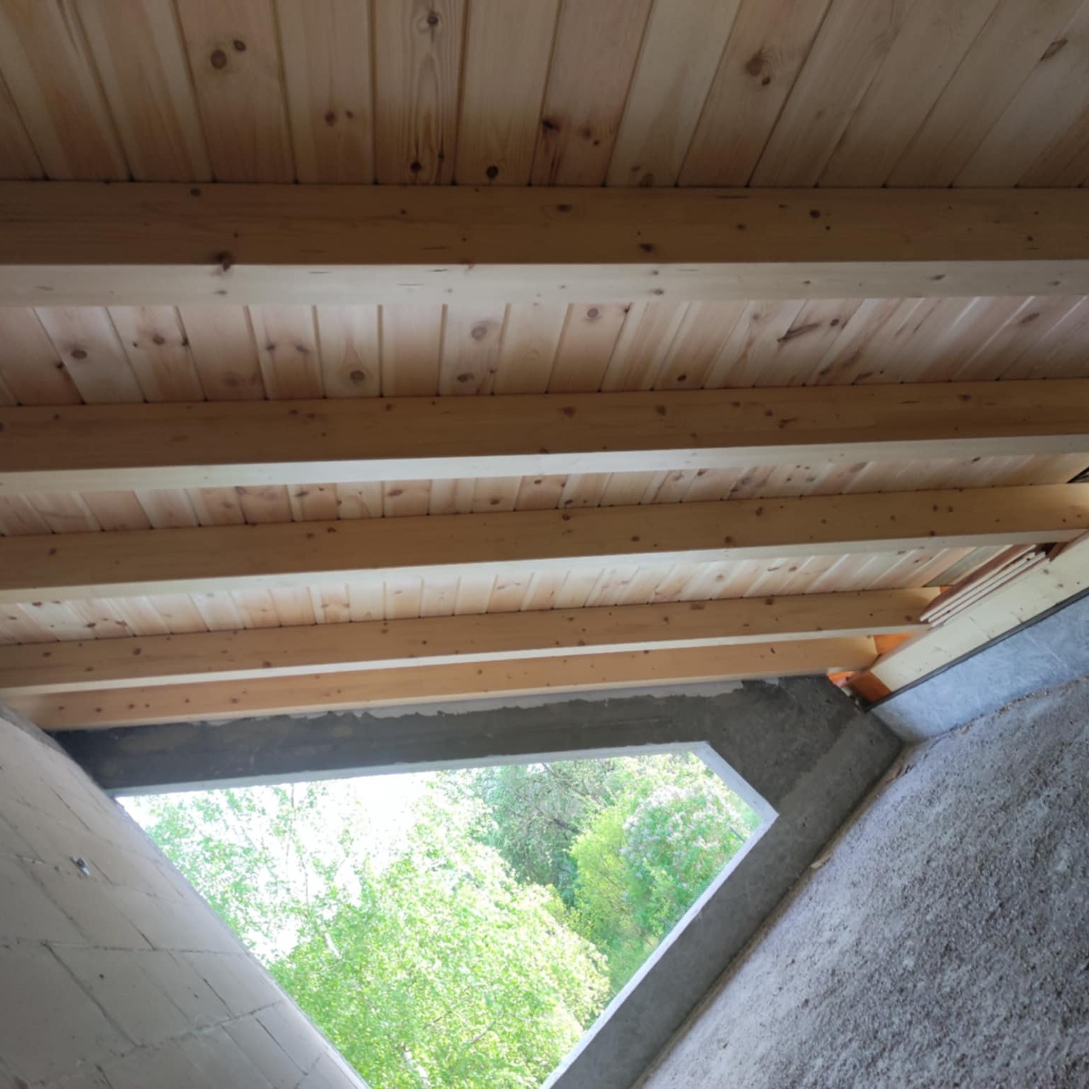 Surowy strop z desek i belek w trakcie budowy, widok z wnętrza przez okno na zieleń. Szare ściany w tle. Ujęcie z dołu, podkreślające konstrukcję.