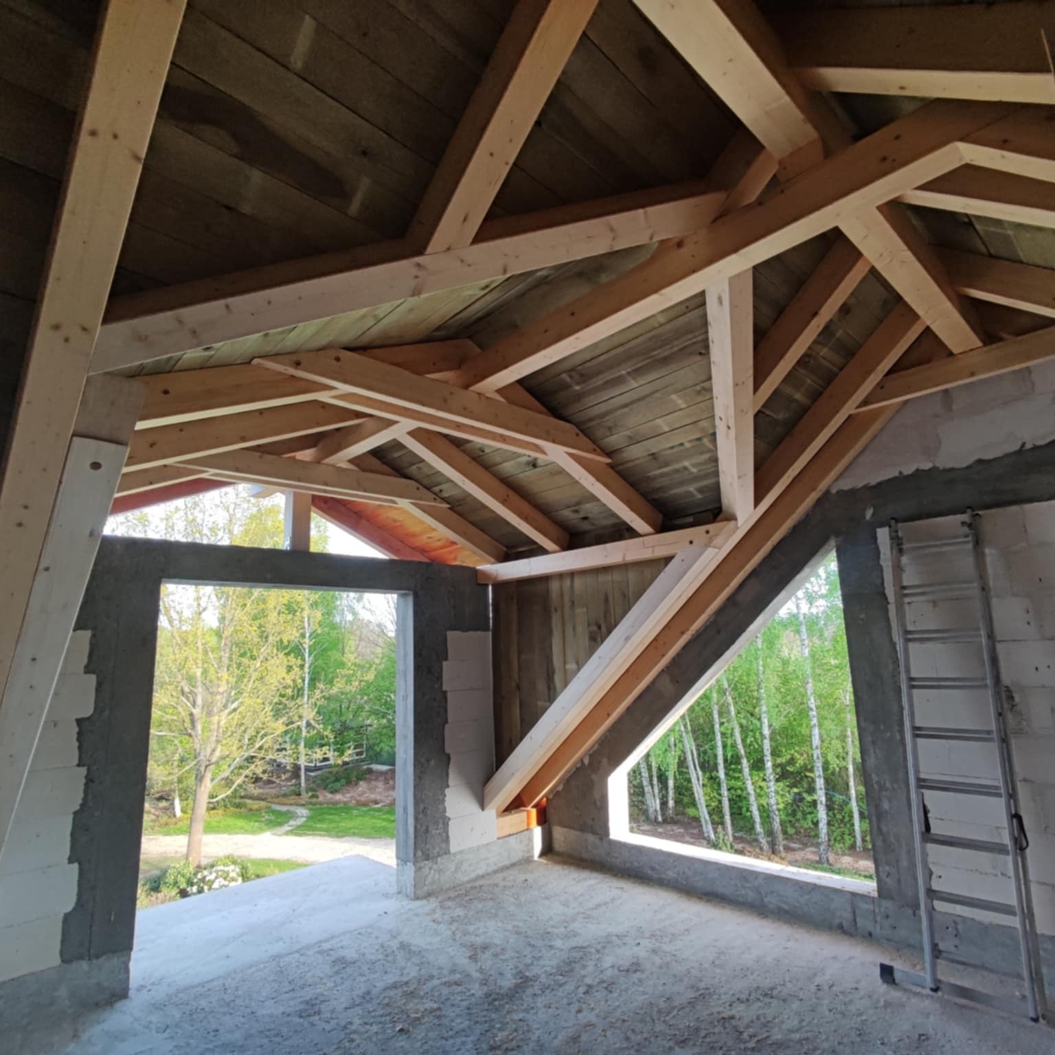 Wnętrze w trakcie budowy z widoczną drewnianą konstrukcją dachu i surowymi ścianami. W tle zieleń drzew widoczna przez otwory okienne. Metalowa drabina oparta o ścianę.