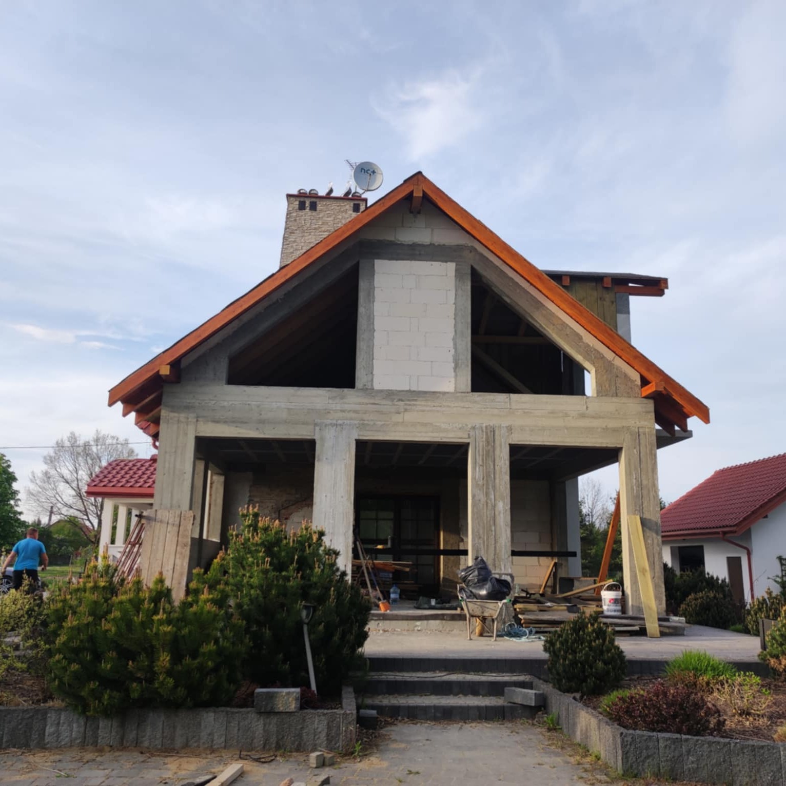 Surowy stan domu w budowie z widocznym szkieletem konstrukcyjnym, kominem i elementami wykończeniowymi dachu. Na pierwszym planie roślinność i częściowo widoczny teren budowy.