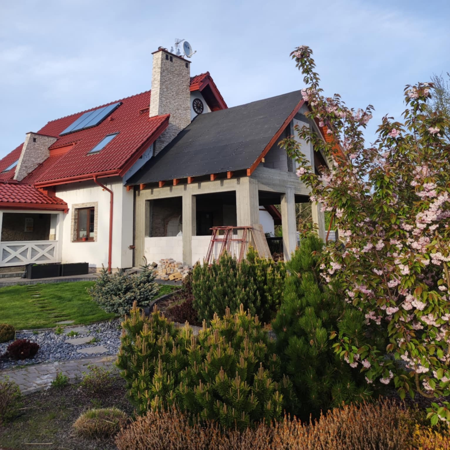 Dom jednorodzinny z czerwoną dachówką i dobudowaną, surową konstrukcją tarasu z czarnym dachem. W tle zielony trawnik i kwitnące drzewo.