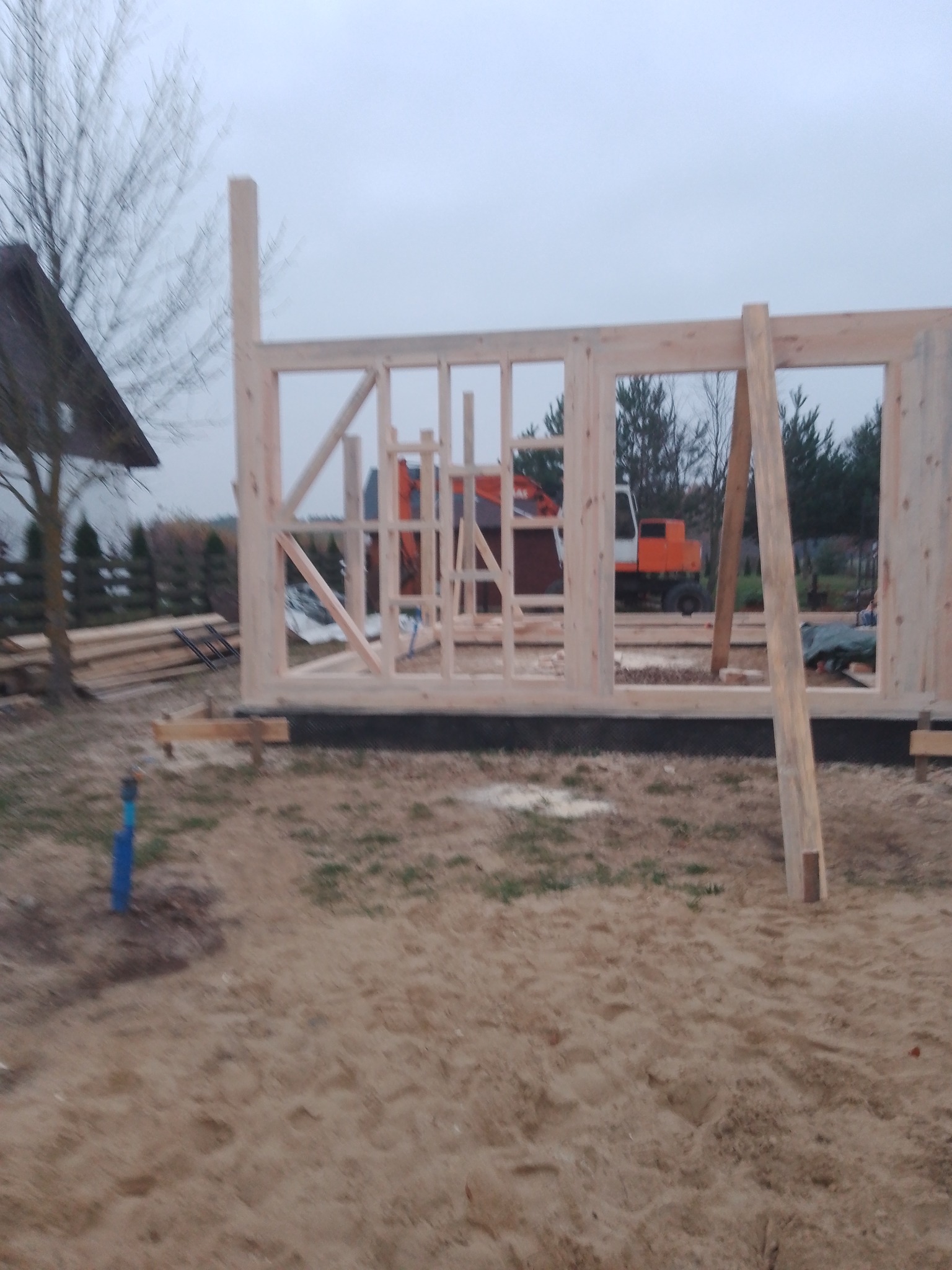 Wznoszona konstrukcja szkieletowa domu z jasnego drewna na fundamencie. W tle widoczny sprzęt budowlany i drzewa. Pochmurne niebo.